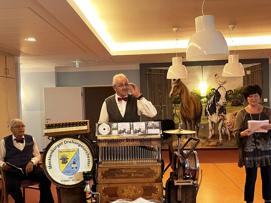 Drei Personen stehen in einem Raum. Zwei ältere Männer in weißem Hemd, Weste und Fliege stehen neben einer Drehorgel. Eine Frau mit dunklen Haaren hält ein Buch oder Notiz in der Hand.