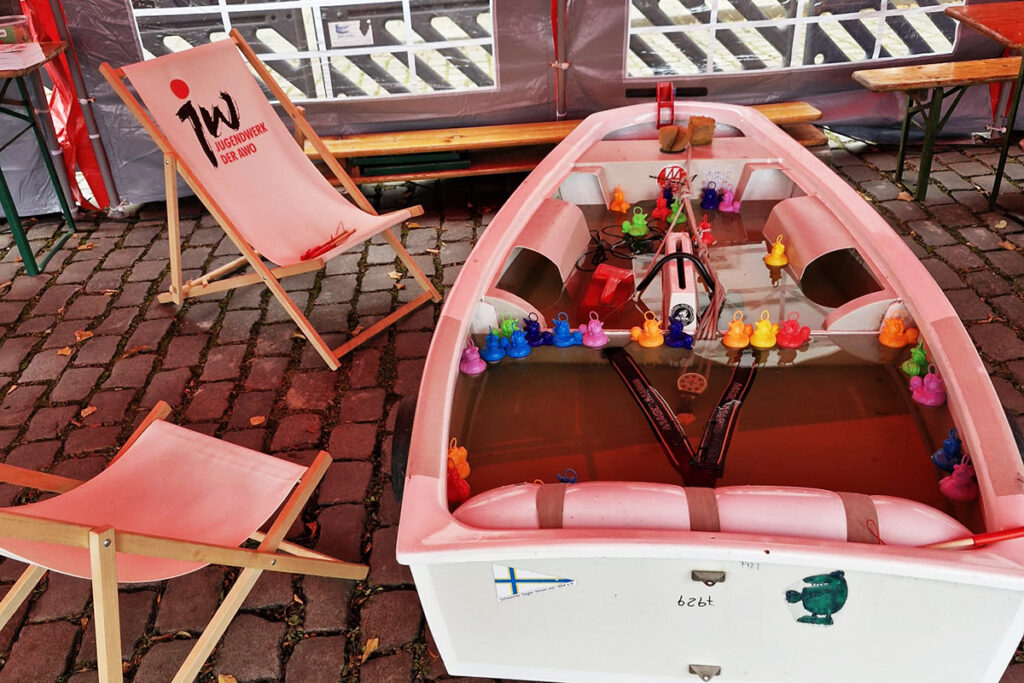 Ein kleines Spielzeugboot gef&uuml;llt mit Wasser, umgeben von bunten Gummienten, steht auf einem gepflasterten Boden neben zwei Liegest&uuml;hlen.