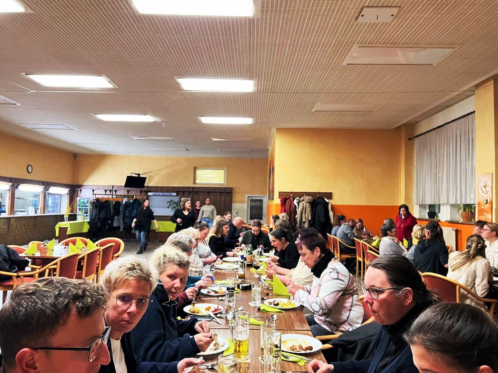 Menschen sitzen in einem großen Raum an einem langen Tisch und essen gemeinsam.
