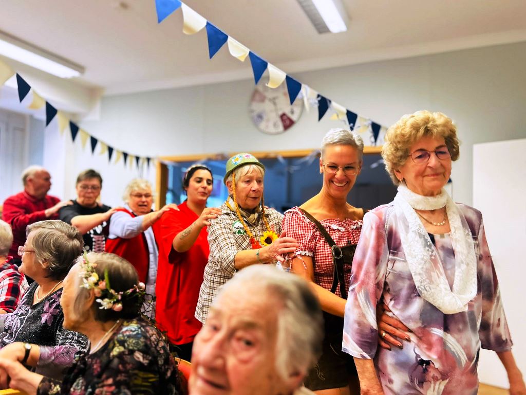 Eine Gruppe älterer Menschen, die fröhlich aneinander gereiht und tanzend eine Polonaise in einem festlich geschmückten Raum machen.