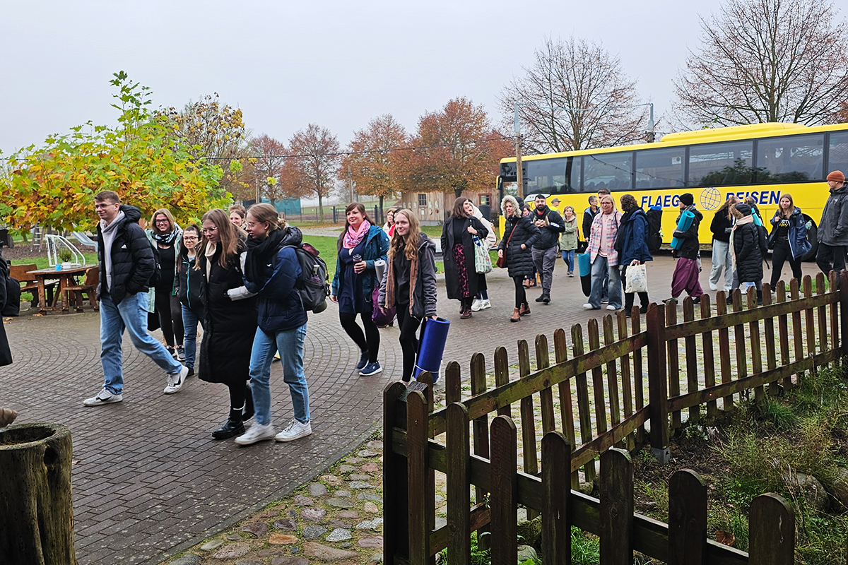 Eine Gruppe von Menschen geht eine gepflasterte Straße entlang, im Hintergrund ein gelber Bus und herbstliche Bäume.