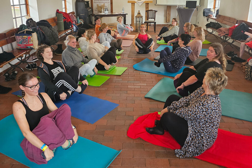 Menschen sitzen in einem Kreis auf bunten Yogamatten in einem Raum.