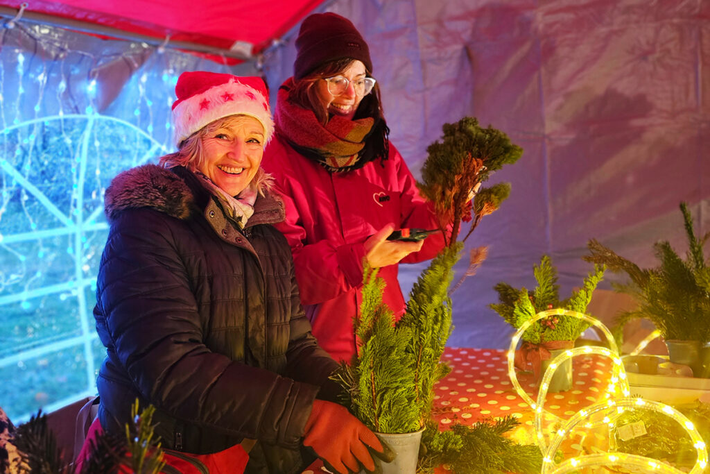 Zwei Personen stehen an einem festlich geschm&uuml;ckten Tisch, umgeben von Lichtern und kleinen Tannenb&auml;umen.