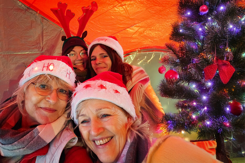 Vier Personen mit roten Weihnachtsm&uuml;tzen, lachend neben einem festlich geschm&uuml;ckten Weihnachtsbaum.