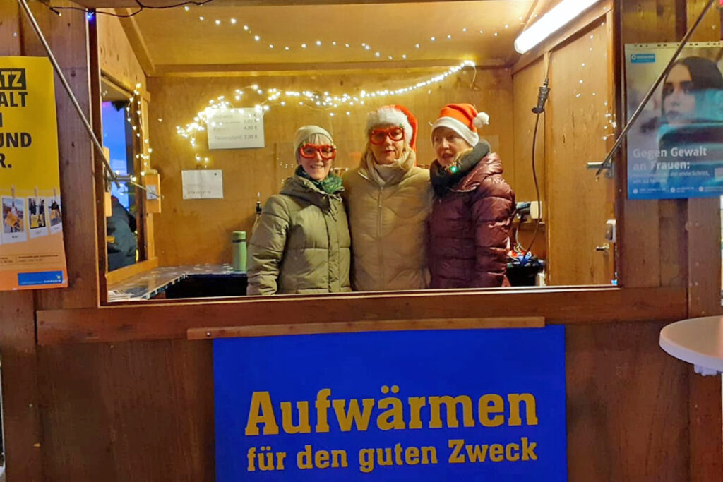 Drei Personen stehen in einer h&ouml;lzernen Verkaufsh&uuml;tte, geschm&uuml;ckt mit Lichterketten. Sie tragen weihnachtliche M&uuml;tzen und rote Brillen. Vor der H&uuml;tte ist ein blaues Schild mit der Aufschrift 'Aufw&auml;rmen f&uuml;r den guten Zweck'.