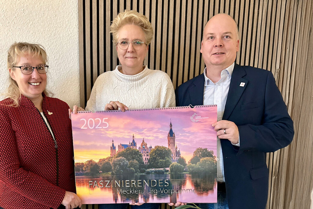 Drei Personen halten einen Kalender für 2025 mit einem Bild von Schloss Schwerin.