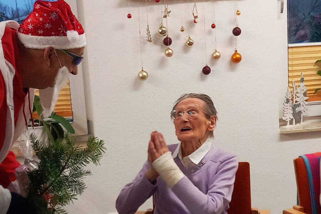 &Auml;ltere Frau in einem Raum l&auml;chelt zu einem als Weihnachtsmann verkleideten Mann hoch.