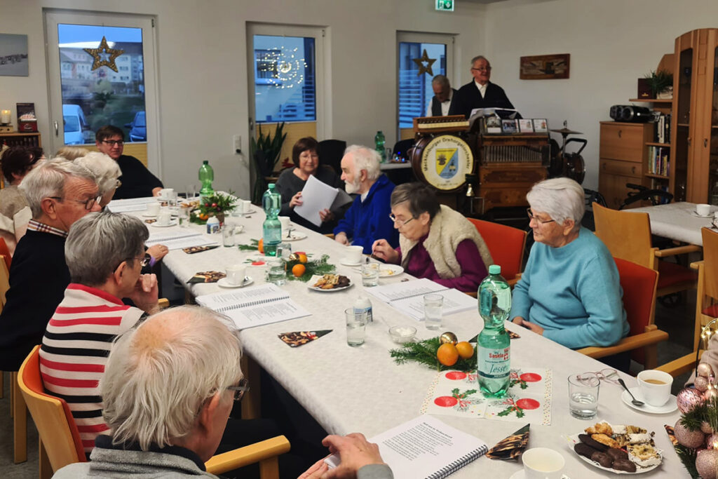 Eine Gruppe &auml;lterer Menschen sitzt an einem Tisch und isst und trinkt gemeinsam. Im Hintergrund spielt jemand auf einer Drehorgel.