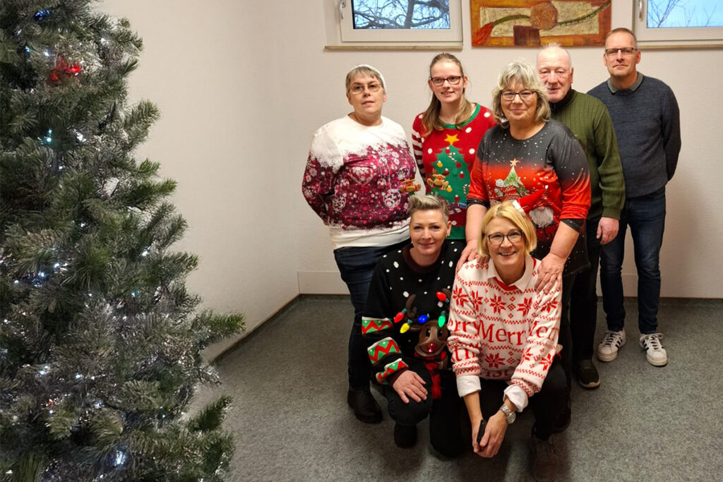 Eine Gruppe von sieben Personen posiert in weihnachtlichen Pullovern neben einem geschm&uuml;ckten Weihnachtsbaum.