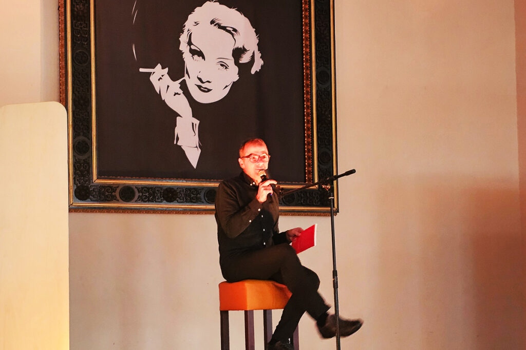 Mann sitzt auf einem Stuhl vor einem gro&szlig;en Portr&auml;trahmen und h&auml;lt ein Mikrofon.