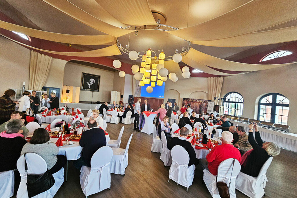 Festlich gedeckter Veranstaltungssaal mit Menschen an Tischgruppen.