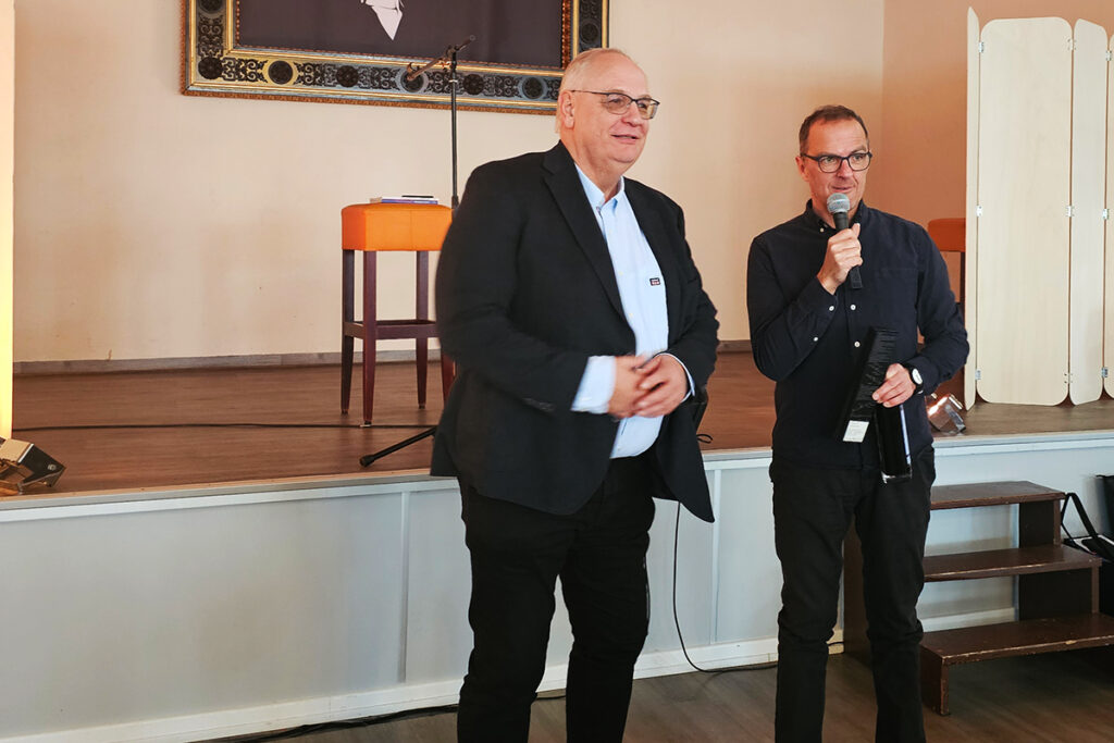 Zwei M&auml;nner stehen auf einer B&uuml;hne; einer h&auml;lt ein Mikrofon.