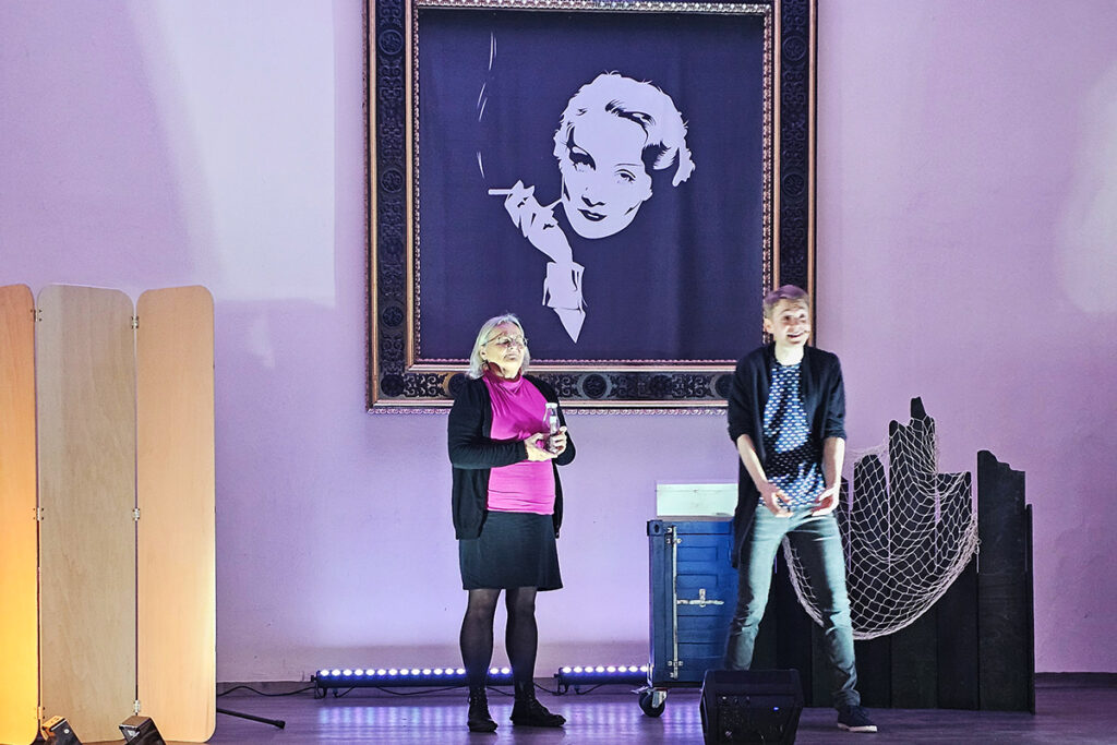 Zwei Personen auf einer B&uuml;hne mit gro&szlig;em Kunstwerksrahmen an der Wand