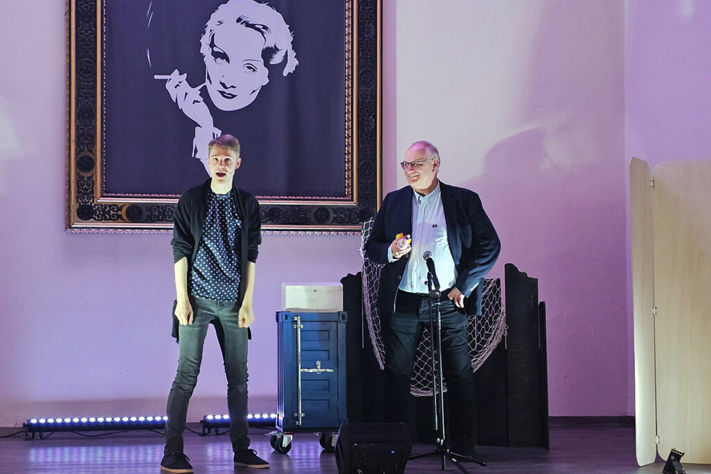 Eine Szene w&auml;hrend einer Veranstaltung mit zwei M&auml;nnern auf einer B&uuml;hne unter einem gro&szlig;en Bild.