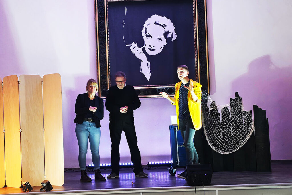 Drei Personen auf einer B&uuml;hne mit einem gro&szlig;en Portr&auml;t im Hintergrund.