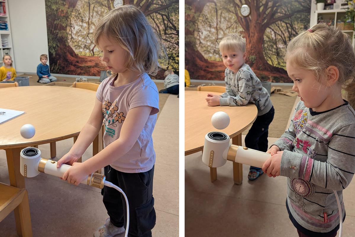 Zwei Kinder experimentieren mit einem Haartrockner und einem Ball in einem Raum mit Baumtapete im Hintergrund.