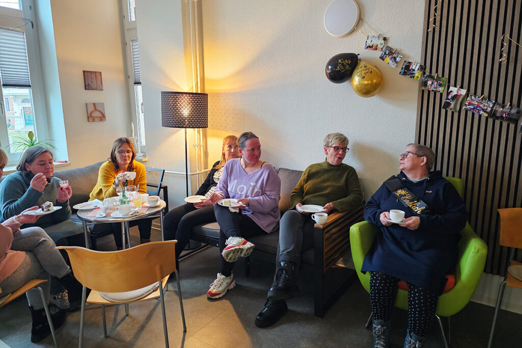 Menschen sitzen in einem Wohnzimmer und unterhalten sich bei Kaffee und Kuchen.