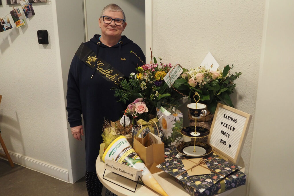 Eine l&auml;chelnde Person steht neben einem Tisch mit Geschenken und Blumen.