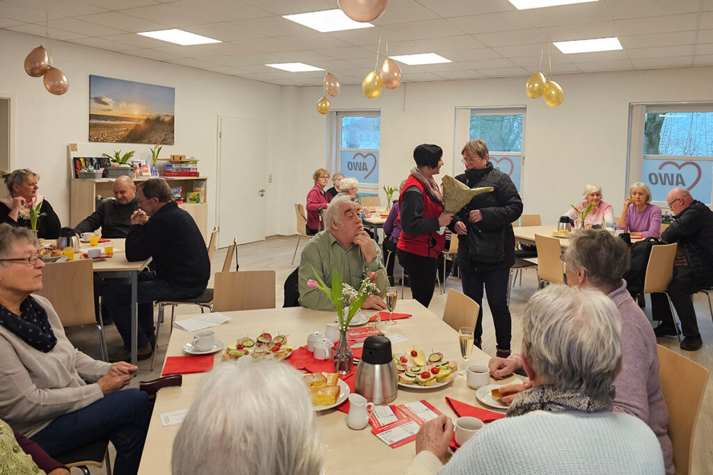 Eine Gruppe von Menschen sitzt in einem freundlich eingerichteten Raum an Tischen und genie&szlig;t eine Mahlzeit.