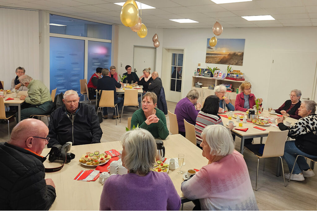 Eine Gruppe von Menschen sitzt in einem Gemeinschaftsraum an Tischen und unterh&auml;lt sich.
