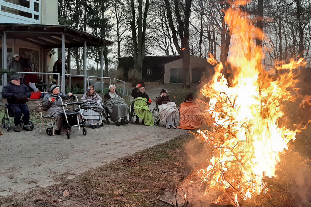 Menschen in Rollst&uuml;hlen und auf B&auml;nken sitzen vor einem Feuer.