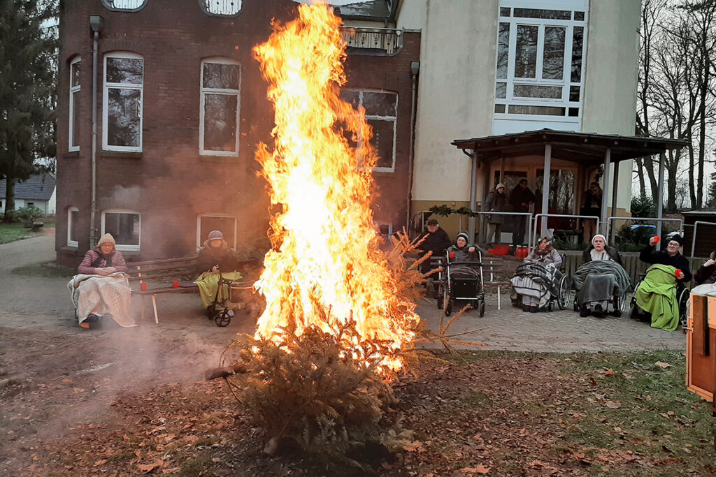 Menschen sitzen in Rollst&uuml;hlen vor einem Geb&auml;ude und beobachten ein gro&szlig;es Feuer.