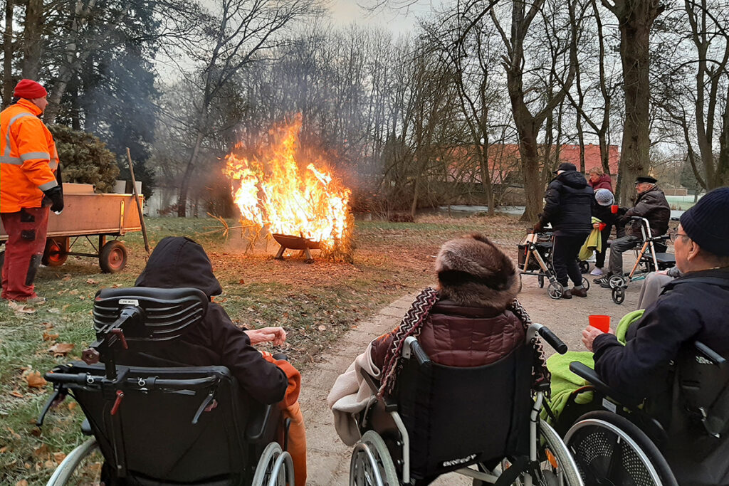 Mehrere Menschen, einige in Rollst&uuml;hlen, sitzen und schauen auf ein brennendes Feuer im Freien.