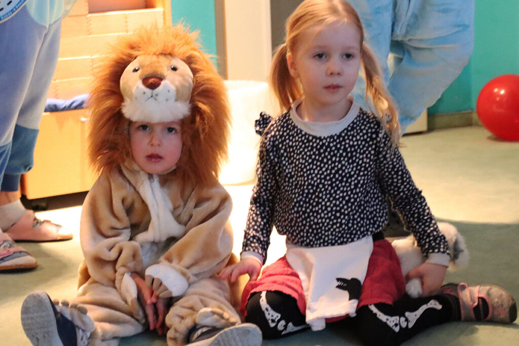 Zwei kleine Kinder sitzen nebeneinander auf dem Boden, eines als Löwe verkleidet, das andere in einem gepunkteten Oberteil mit Zöpfen.