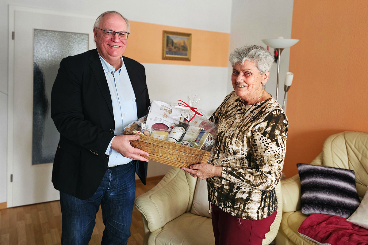 Ein älterer Mann und eine ältere Frau halten gemeinsam einen Geschenkekorb in einem Wohnzimmer.