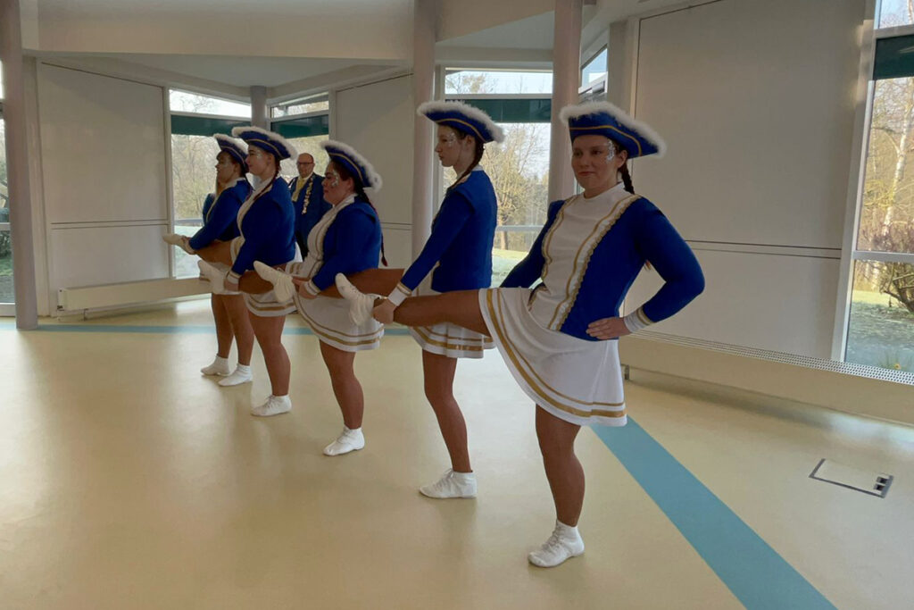 Gruppe von T&auml;nzerinnen in blauen Kost&uuml;men mit wei&szlig;en R&ouml;cken und H&uuml;ten, die in einer Reihe stehen und einen Tritt ausf&uuml;hren.
