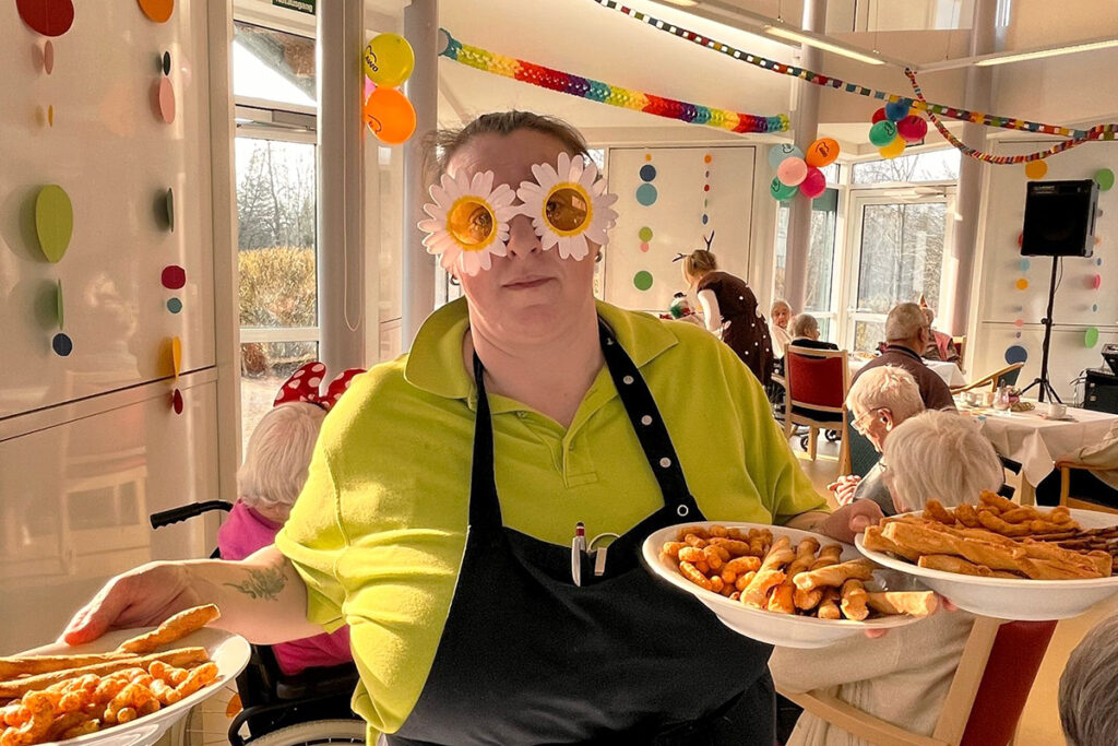 Person mit Sonnenblumenbrille tr&auml;gt zwei Teller mit Snacks in einem festlich dekorierten Raum.