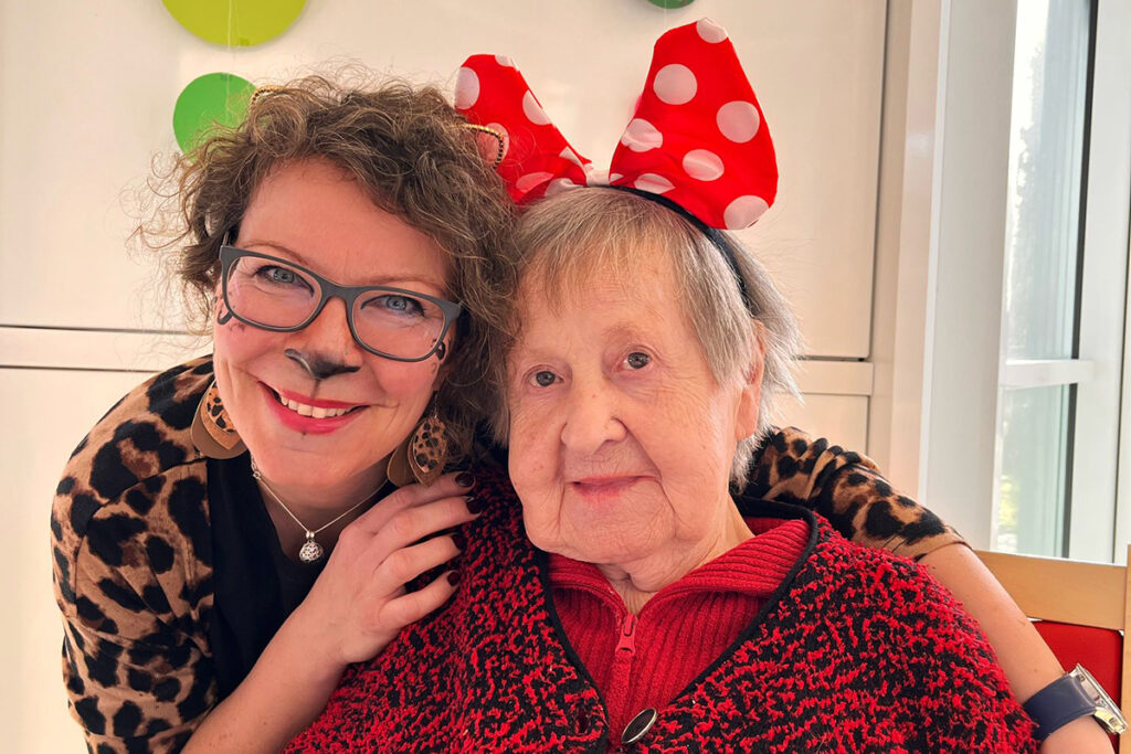 Zwei Frauen in festlicher Kleidung, eine &auml;ltere und eine j&uuml;ngere, die in die Kamera l&auml;cheln, die j&uuml;ngere Frau tr&auml;gt Tierohren und Gesichtsbemalung, die &auml;ltere Frau tr&auml;gt eine rote Strickjacke.