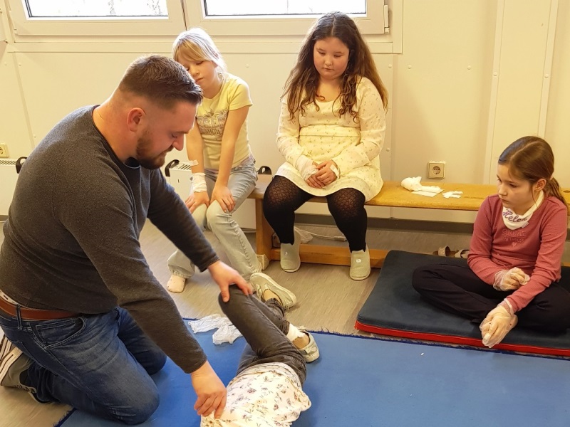 Ein Mann demonstriert eine Erste-Hilfe-Technik an einem Kind, während drei Kinder zuschauen.