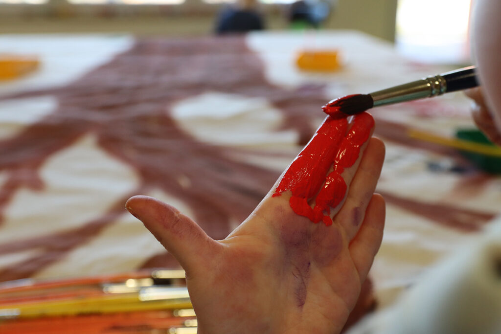 Nahaufnahme einer Kinderhand mit roter Farbe bemalt, ein Pinsel in der anderen Hand
