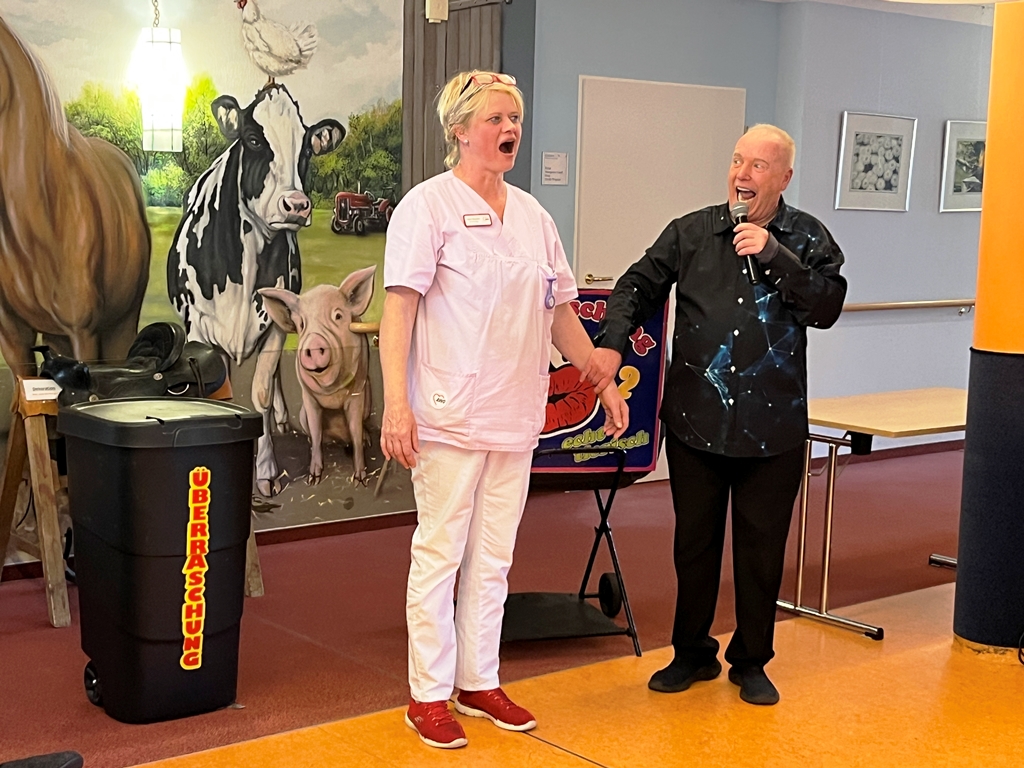 Eine Person in einem weißen Pflegeoutfit steht lächelnd neben einer Person mit Mikrofon, die in ein schwarzes Hemd gekleidet ist. Im Hintergrund ist eine Wand mit einem Tiermotiv bemalt.