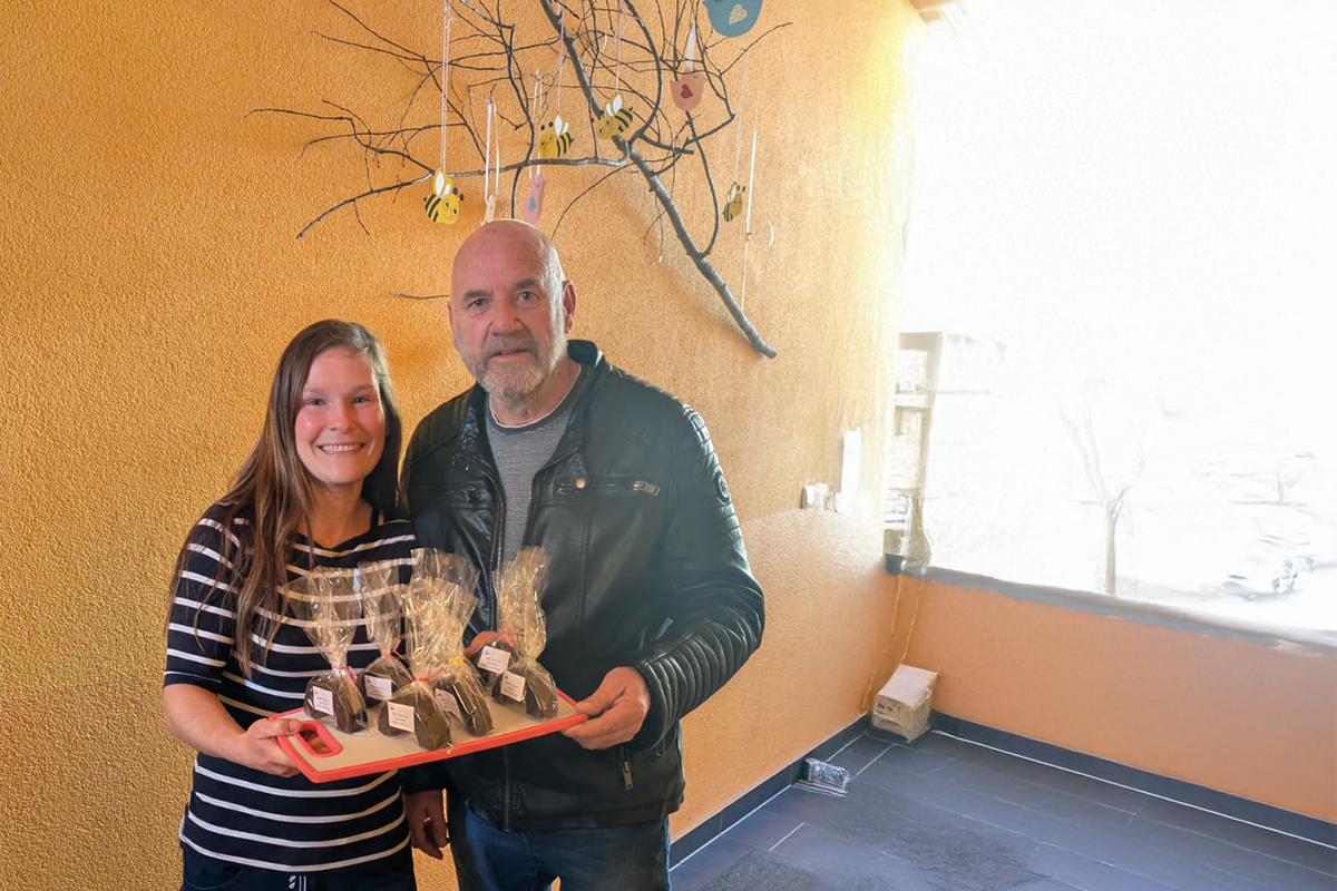 Eine Frau und ein Mann halten ein Tablett mit verpackten Produkten vor einem gelben Hintergrund.