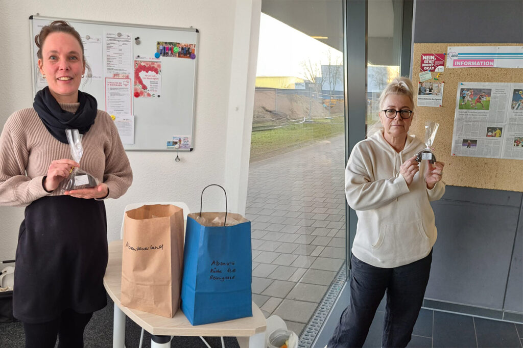 Zwei Frauen halten kleine P&auml;ckchen in einem Raum mit einer Pinnwand.