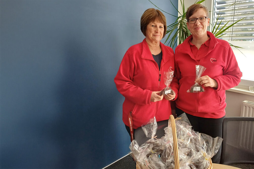 Zwei Personen in roten AWO-Jacken halten kleine cellophanverpackte Geschenke in den H&auml;nden. Vor ihnen steht ein Korb mit weiteren Geschenken.