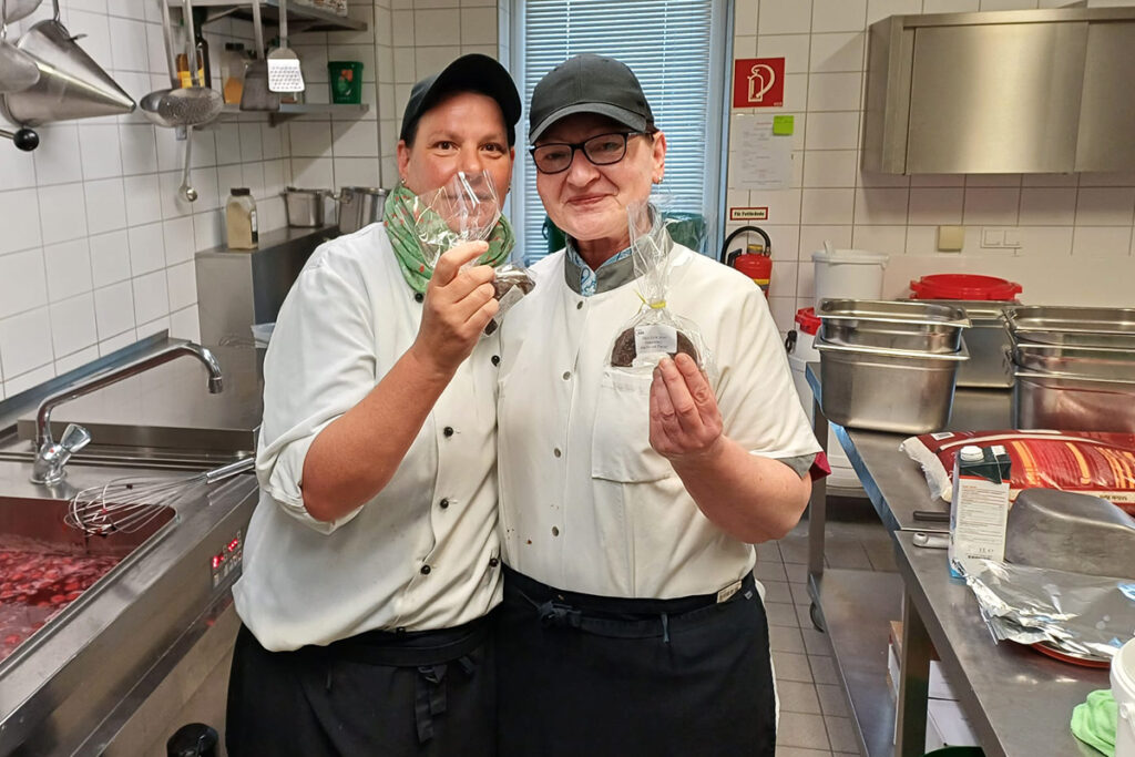 Zwei K&uuml;chenmitarbeiterinnen in einer Gro&szlig;k&uuml;che l&auml;cheln in die Kamera und halten verpackte Lebensmittel in den H&auml;nden.