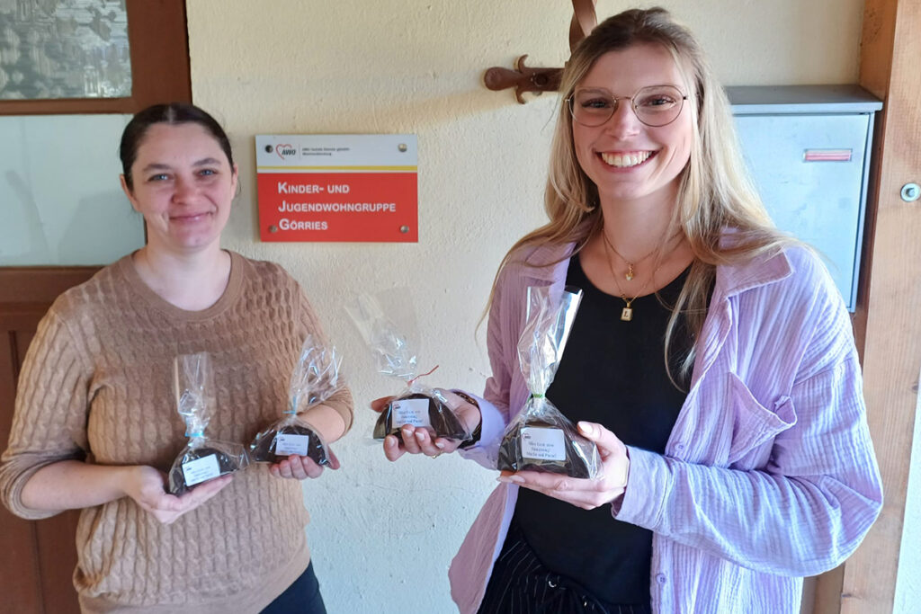 Zwei Frauen stehen mit Geschenkt&uuml;ten in den H&auml;nden vor einem Schild mit der Aufschrift 'Kinder- und Jugendwohngruppe Gorries'.