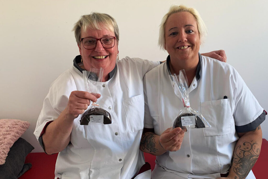 Zwei lachende Personen in wei&szlig;en Kitteln halten in Klarsichtfolie verpackte kleine Kuchen.