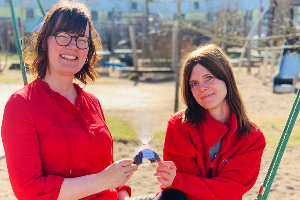 Zwei Frauen in roten Oberteilen auf einem Spielplatz, die ein verpacktes Geschenk halten.