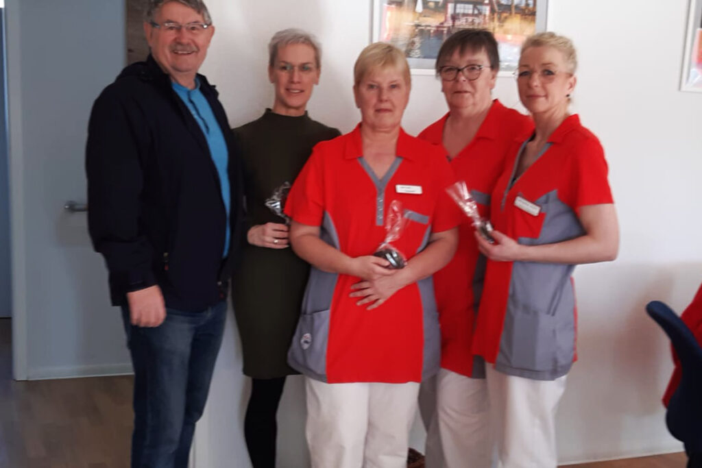 Eine Gruppe von f&uuml;nf Menschen, darunter vier Frauen in roten Uniformen und ein Mann in Freizeitkleidung, die l&auml;chelnd zusammenstehen.