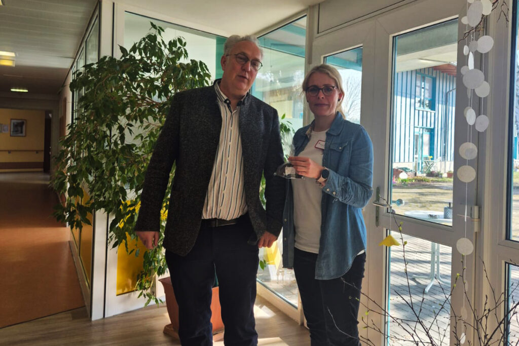 Zwei Personen stehen in einem lichtdurchfluteten Raum neben einem gro&szlig;en Fenster.