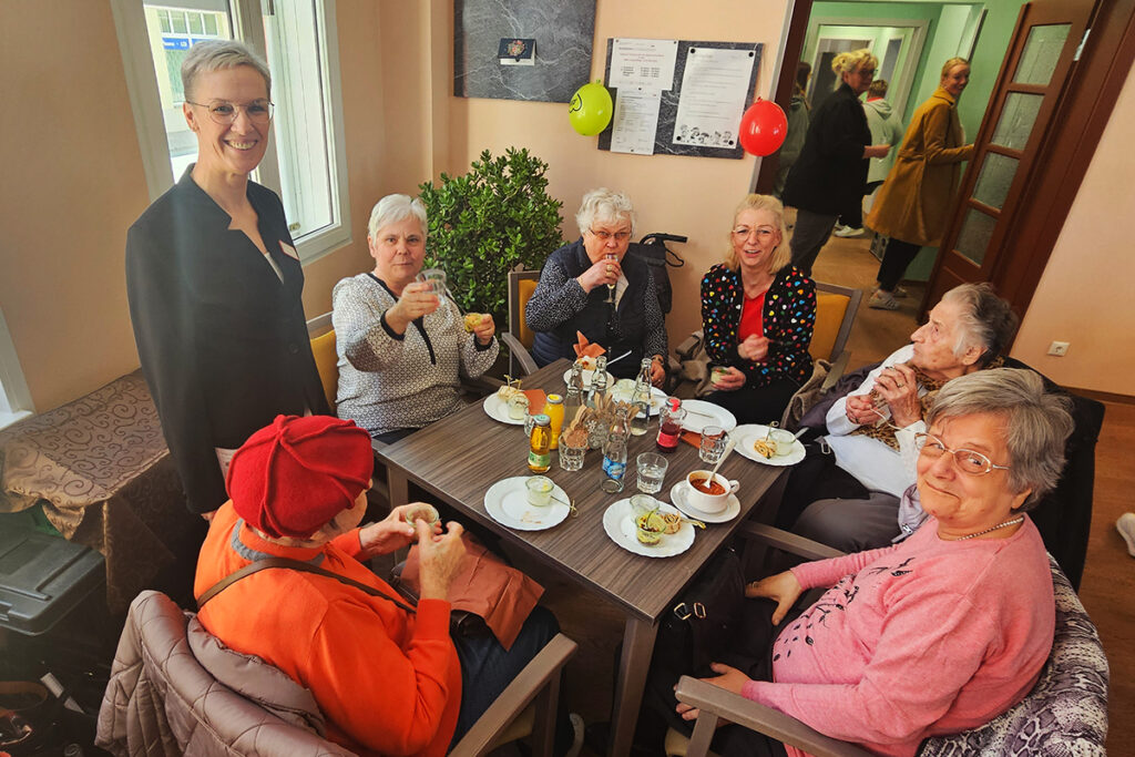Gruppenszene in einem Raum, in dem mehrere &auml;ltere Frauen um einen Tisch sitzen und eine Frau steht.
