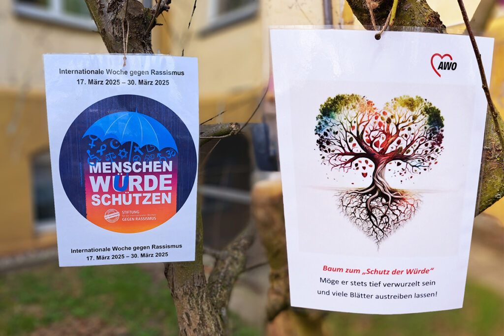 Zwei Plakate an einem Baum, das linke mit einem blauen Regenschirm und Text 'Menschenw&uuml;rde sch&uuml;tzen', das rechte mit einem gezeichneten Baum in Herzform.