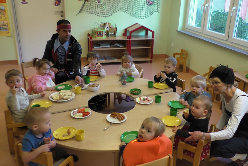 Kindergartenkinder sitzen an einem runden Tisch und essen zusammen mit zwei Erwachsenen, die sich verkleidet haben.