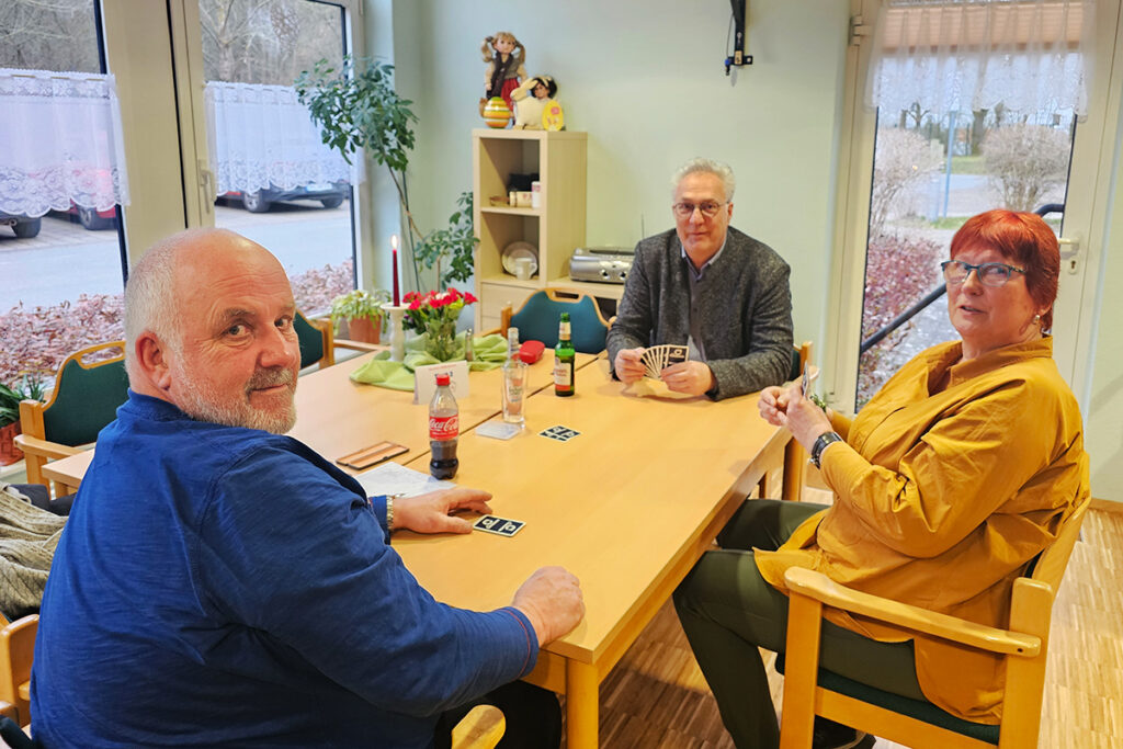 Drei Personen sitzen an einem Tisch und spielen ein Kartenspiel.