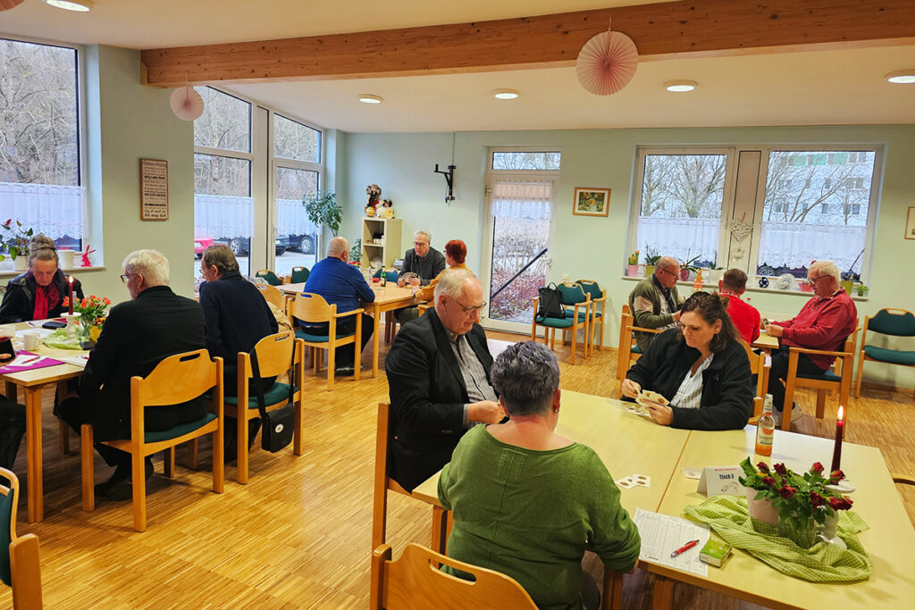 Menschen in einem Gemeinschaftsraum sitzen an Tischen und spielen Karten.