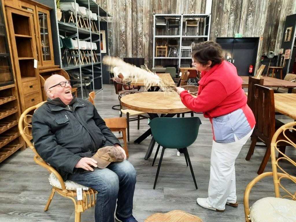 Zwei Personen in einem Möbelgeschäft, eine Person sitzt lachend auf einem Stuhl, während die andere sie spielerisch mit einem Bündel Stroh kitzelt.